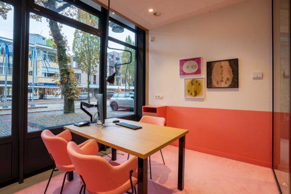 Modern ingericht kantoor met roze accenten, een bureau met vier stoelen en een groot raam met uitzicht op de 1e Hogeweg.